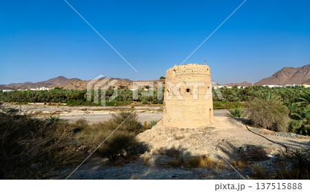 The historic stone structure of Al Basra Fort Tower stands near a lush palm oasis and dry riverbed in the scenic mountain landscape of Fanja, Oman. The historic stone structure of Al Basra Fort Tower stands near a lush palm oasis and dry riverbed in the scenic mountain landscape of Fanja, Oman. 137515888