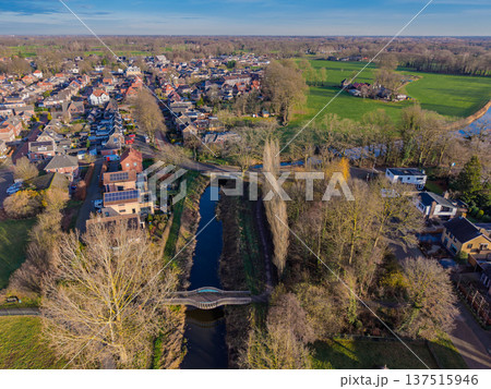 Aerial view of a Dutch town featuring a canal with a footbridge, residential houses, and green meadows. Scenic suburban landscape in the Netherlands during a sunny autumn day. Aerial view of a Dutch town featuring a canal with a footbridge, residential houses, and green meadows. Scenic suburban landscape in the Netherlands during a sunny autumn day. 137515946