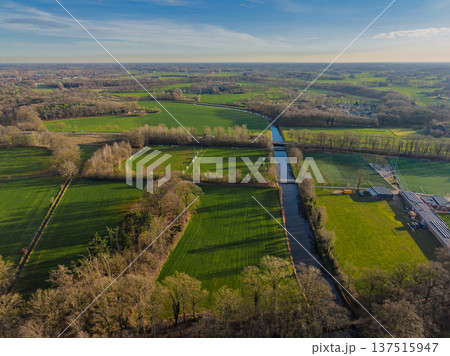 Aerial view of Dutch countryside with a long canal, soccer fields, and green meadows. Scenic rural landscape with trees and distant village under a clear sky at sunset. 137515947