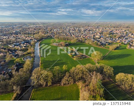 Wide aerial panorama of a Dutch town border with a winding canal, green meadows, and residential districts. Scenic European rural urban landscape under a blue sky at sunset. Wide aerial panorama of a Dutch town border with a winding canal, green meadows, and residential districts. Scenic European rural urban landscape under a blue sky at sunset. 137515948