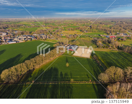 High aerial view of a Dutch landscape featuring horse stables, green pastures, and a distant town with wind turbines. Scenic rural countryside in the Netherlands during late afternoon sun. 137515949