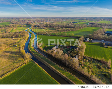 Aerial view of a scenic Dutch landscape with a long canal cutting through vibrant green fields and meadows. Rural countryside in the Netherlands under a clear blue sky at sunset. Aerial view of a scenic Dutch landscape with a long canal cutting through vibrant green fields and meadows. Rural countryside in the Netherlands under a clear blue sky at sunset. 137515952