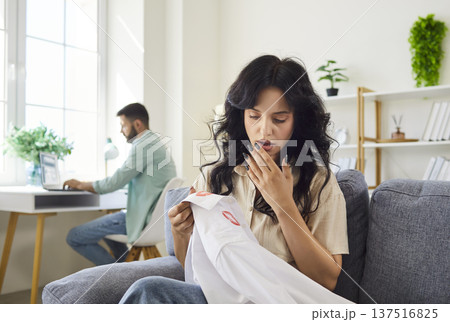 Jealous wife in shock and stress holding husbands shirt with lipstick marks Jealous wife in shock and stress holding husbands shirt with lipstick marks 137516825