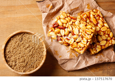 A piece of Brazilian peanut brittle made with roasted peanuts and golden caramel, on kraft paper on a rustic wooden table next to a bowl of sugar A piece of Brazilian peanut brittle made with roasted peanuts and golden caramel, on kraft paper on a rustic wooden table next to a bowl of sugar 137517670