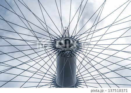 Central steel hub and cable spokes of a large observation wheel photographed from below, showing geometric symmetry and modern engineering structure against a cloudy sky. Central steel hub and cable spokes of a large observation wheel photographed from below, showing geometric symmetry and modern engineering structure against a cloudy sky. 137518371