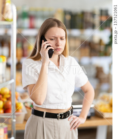 Upset young woman choosing groceries while talking on phone Upset young woman choosing groceries while talking on phone 137521061