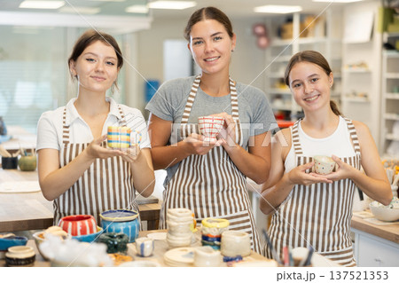 Three positive girls stands in pottery workshop and show created work, finished decorative vase 137521353
