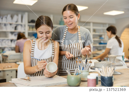 During master class, teen girl student paints clay vessel under guidance of female teacher. 137521369