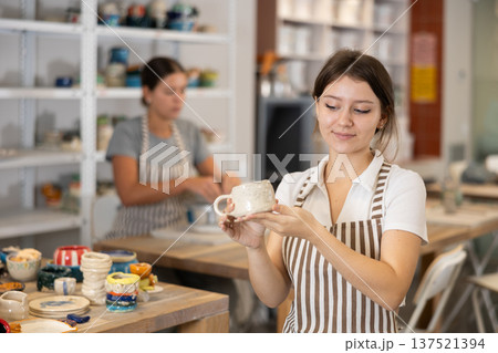 Girl stands in pottery workshop and shows created work, finished decorative vase - jug. 137521394