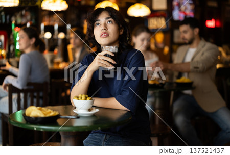 Glad young girl relaxing at table and drinking beer in sports pub, attentively watching match on TV Glad young girl relaxing at table and drinking beer in sports pub, attentively watching match on TV 137521437
