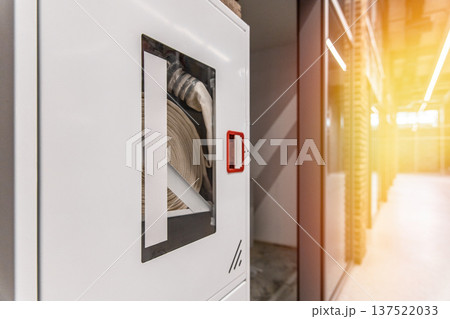 Fire safety equipment in a modern office. White hydrant cabinet with a fire hose behind glass in an industrial interior corridor. 137522033