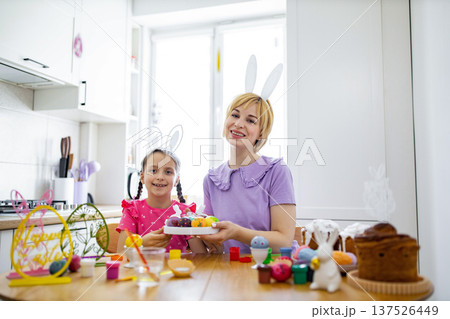 A mother and daughter wearing bunny ears are happily painting Easter eggs together in a bright kitchen setting 137526449
