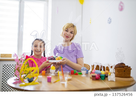 A mother and daughter wearing bunny ears joyfully prepare for Easter by decorating colorful eggs and baking traditional cakes together 137526456