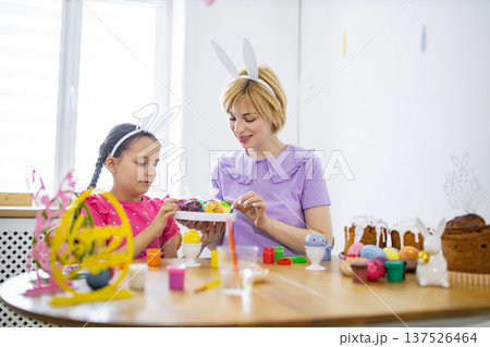 A mother and her daughter wearing bunny ears are happily decorating colorful Easter eggs together at a wooden table filled with festive treats and craft supplies 137526464