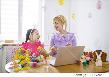 A mother and daughter wearing bunny ears are painting Easter eggs together at a table with a laptop and Easter cakes A mother and daughter wearing bunny ears are painting Easter eggs together at a table with a laptop and Easter cakes 137526468