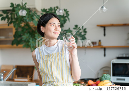 キッチンで水分補給をするエプロン姿の若いアジア人女性 キッチンで水分補給をするエプロン姿の若いアジア人女性 137526716