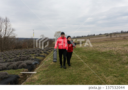 Two individuals carrying rolled mats in a lavender field under a cloudy sky during the cool hours of early spring in a serene rural landscape 137532044