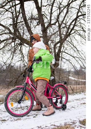 Joyful winter adventure as a young boy rides his bright pink bicycle while his friend lends a helping hand under a tall, leafless tree in the snowy landscape Joyful winter adventure as a young boy rides his bright pink bicycle while his friend lends a helping hand under a tall, leafless tree in the snowy landscape 137532245