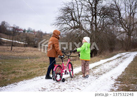 Two children chatting on a snowy path while one holds a pink bicycle, set in a serene winter landscape with patches of snow and overcast skies in a suburban area 137532248