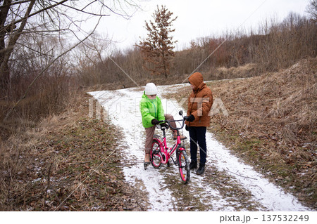 Two children enjoying a winter day on a snowy path while one inspects a pink bicycle amidst a serene nature backdrop in a quiet outdoor location Two children enjoying a winter day on a snowy path while one inspects a pink bicycle amidst a serene nature backdrop in a quiet outdoor location 137532249