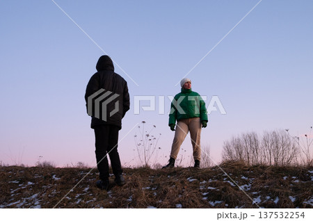 Two figures on a winter evening hillside embracing the cool breeze while the sky transitions to twilight in a serene landscape Two figures on a winter evening hillside embracing the cool breeze while the sky transitions to twilight in a serene landscape 137532254