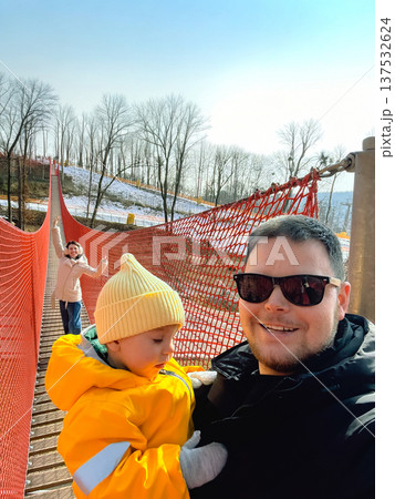 Father, mother and toddler on red rope bridge in winter park Father, mother and toddler on red rope bridge in winter park 137532624