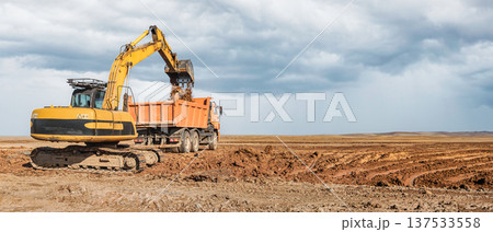 A yellow excavator loads dirt into a large orange truck on a construction site. The site is wide open with dark clouds forming in the sky 137533558