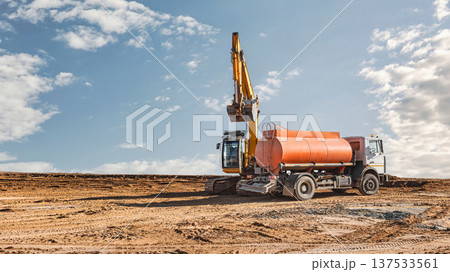 Heavy machinery operates on a dirt construction site under a clear sky. A large truck and an orange tank are positioned for the task at hand Heavy machinery operates on a dirt construction site under a clear sky. A large truck and an orange tank are positioned for the task at hand 137533561