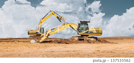 Two excavators work on a construction site, digging and moving dirt under a blue sky with clouds in the background during daytime Two excavators work on a construction site, digging and moving dirt under a blue sky with clouds in the background during daytime 137533562
