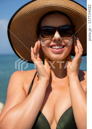Beautiful woman applying sunscreen cream on face while enjoying relaxing beach vacation and protecting skin from UV rays during daily skincare routine 137534082