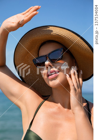 Beautiful woman enjoying sunny beach while applying sunscreen cream on face protecting skin from sunburn using moisturizing suncream lotion during summer skincare routine 137534084