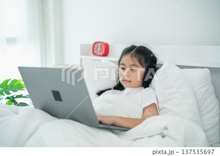 Girl in White Bed Using Laptop with Headphones, Relaxing in Cozy Room, Morning Time, Bright Natural Light, Soft Bedding, Focused Learning Experience 137535697