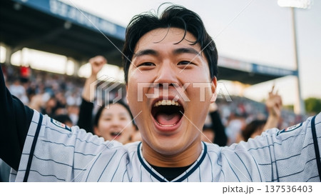 Excited Asian man celebrating victory at baseball stadium wearing striped jersey with arms raised cheering enthusiastically 137536403