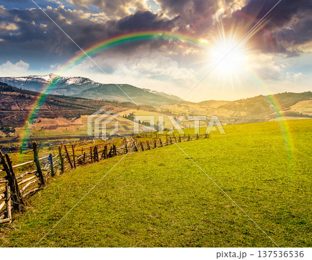 sunset over Springtime rural landscape. Wooden fence along the path through agricultural fields in mountains. beautiful countryside scenery under cloudy sky with rainbow. remote alpine pasture 137536536