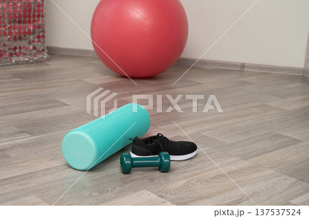 Home workout setup with exercise ball, foam roller, dumbbell, and shoes on wooden floor 137537524