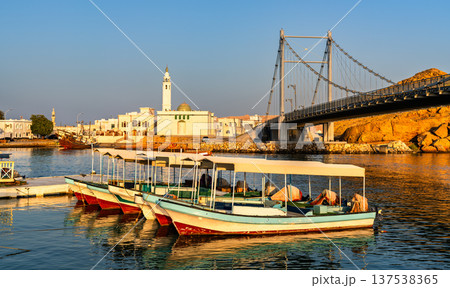 Scenic view of traditional wooden dhow boats moored in the water near the Al Ayjah suspension bridge and a mosque in the coastal city of Sur, Oman at sunset. 137538365