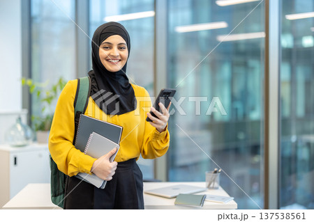 Young muslim woman in a hijab with backpack and notebooks using a smartphone in a bright modern office, smiling and confident while studying or working remotely Young muslim woman in a hijab with backpack and notebooks using a smartphone in a bright modern office, smiling and confident while studying or working remotely 137538561