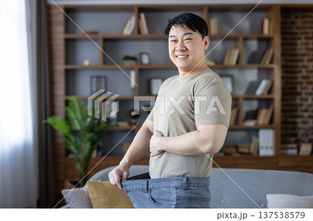 Man celebrating success in weight loss by holding out his baggy oversized jeans, demonstrating the significant transformation and embracing a healthy lifestyle with a cheerful smile at home 137538579