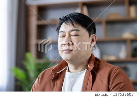 Asian man with closed eyes finding inner balance and feeling relaxed at home, practicing mindfulness, meditation, and mental health for overall well-being Asian man with closed eyes finding inner balance and feeling relaxed at home, practicing mindfulness, meditation, and mental health for overall well-being 137538603