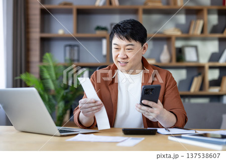 Asian man smiling at home office desk, holding a receipt and using smartphone for online payment and budgeting while working on laptop and organizing finances 137538607