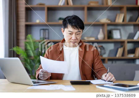 Asian man reviewing documents, writing notes, and calculating expenses, diligently managing personal finances and household budget in his modern home office Asian man reviewing documents, writing notes, and calculating expenses, diligently managing personal finances and household budget in his modern home office 137538609