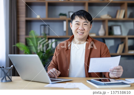 Happy asian man smiling at camera while sitting at his home office desk, holding a stack of papers and working on his laptop, embodying successful remote work and productivity Happy asian man smiling at camera while sitting at his home office desk, holding a stack of papers and working on his laptop, embodying successful remote work and productivity 137538610