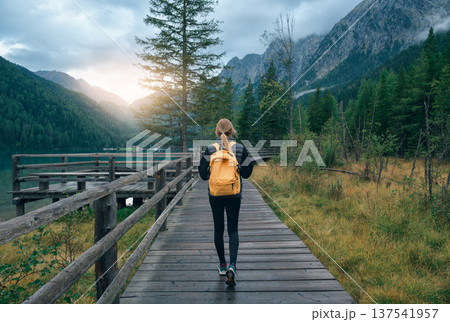 Woman with yellow backpack walking on wooden trail by alpine lak Woman with yellow backpack walking on wooden trail by alpine lak 137541957
