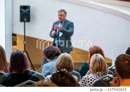 Business speaker addressing audience at indoor conference meeting with professionals listening and taking notes Business speaker addressing audience at indoor conference meeting with professionals listening and taking notes 137542655