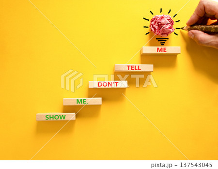 Tell or show symbol. Concept words Show me, do not tell me on beautiful wooden blocks. Beautiful yellow paper background. Light bulb icon. Businessman hand. Business tell or show concept. Copy space. 137543045