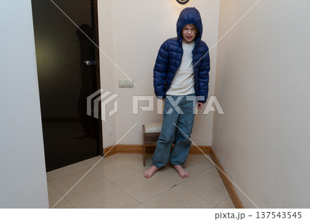 Young barefoot boy in a blue hooded puffer jacket standing indoors. Uncomfortable child grimacing 137543545