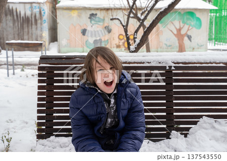 Young boy in blue jacket sitting on a snow-covered bench. Child shouting outdoors in winter Young boy in blue jacket sitting on a snow-covered bench. Child shouting outdoors in winter 137543550