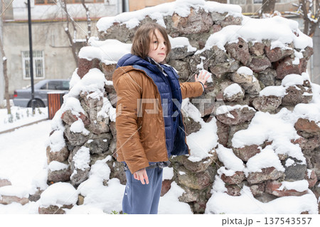 Young boy touching a snow-covered stone wall outdoors. Child exploring a winter urban environment 137543557