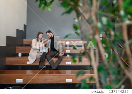 Man and woman talking and holding notebooks surrounded by leafy plants on stepped office bench. Talking, notebooks, plants, teamwork, workspace. Man and woman talking and holding notebooks surrounded by leafy plants on stepped office bench. Talking, notebooks, plants, teamwork, workspace. 137544225