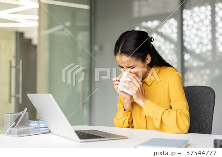 Young biracial woman sitting at her office desk, blowing her nose into a tissue, suffering from a common cold, flu, virus, or seasonal allergy symptoms while working 137544777